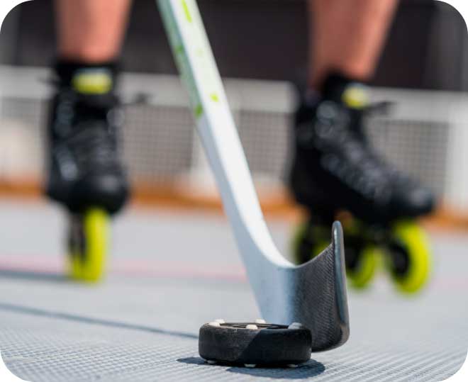 A close-up image of the blade of a hockey stick with 3-wheel inline hockey skates in the background
